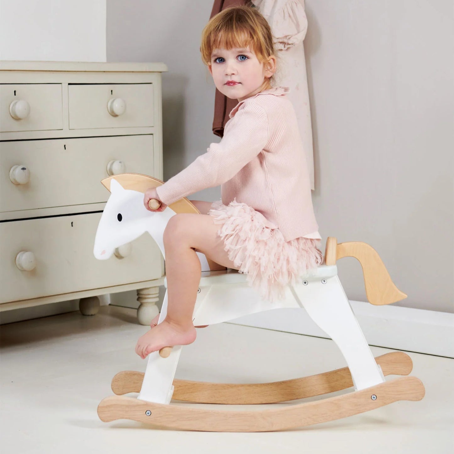 Child sitting on a wooden rocking horse in a room with a dresser and hanging clothes.
