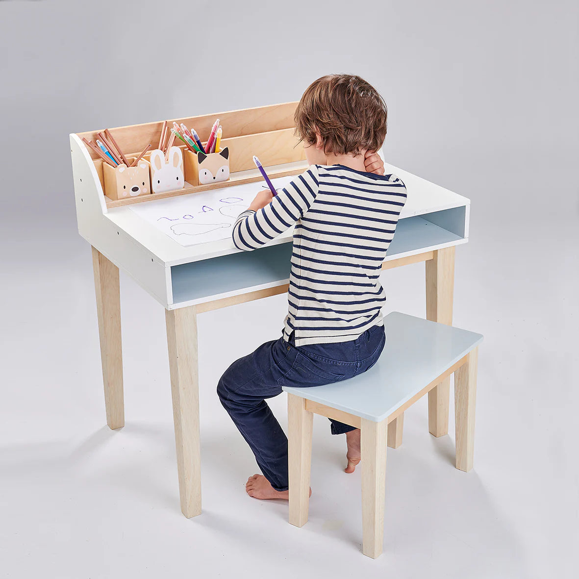 Child sitting at a small desk with a stool, drawing with pencils on a white background