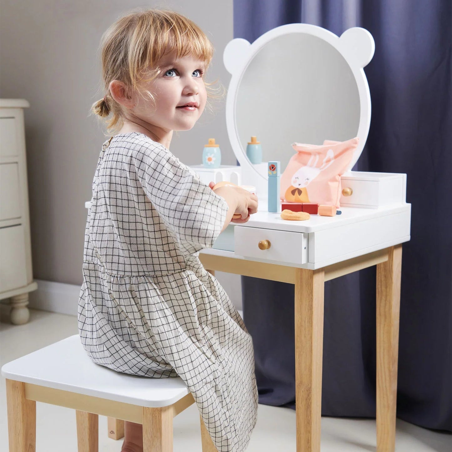 Child sitting at a small vanity table with mirror and toys in a room.