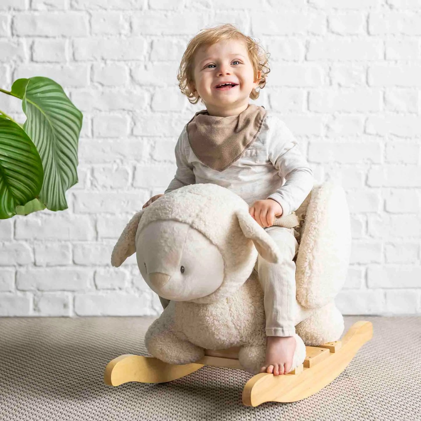 Child sitting on a rocking sheep toy against a white brick wall.