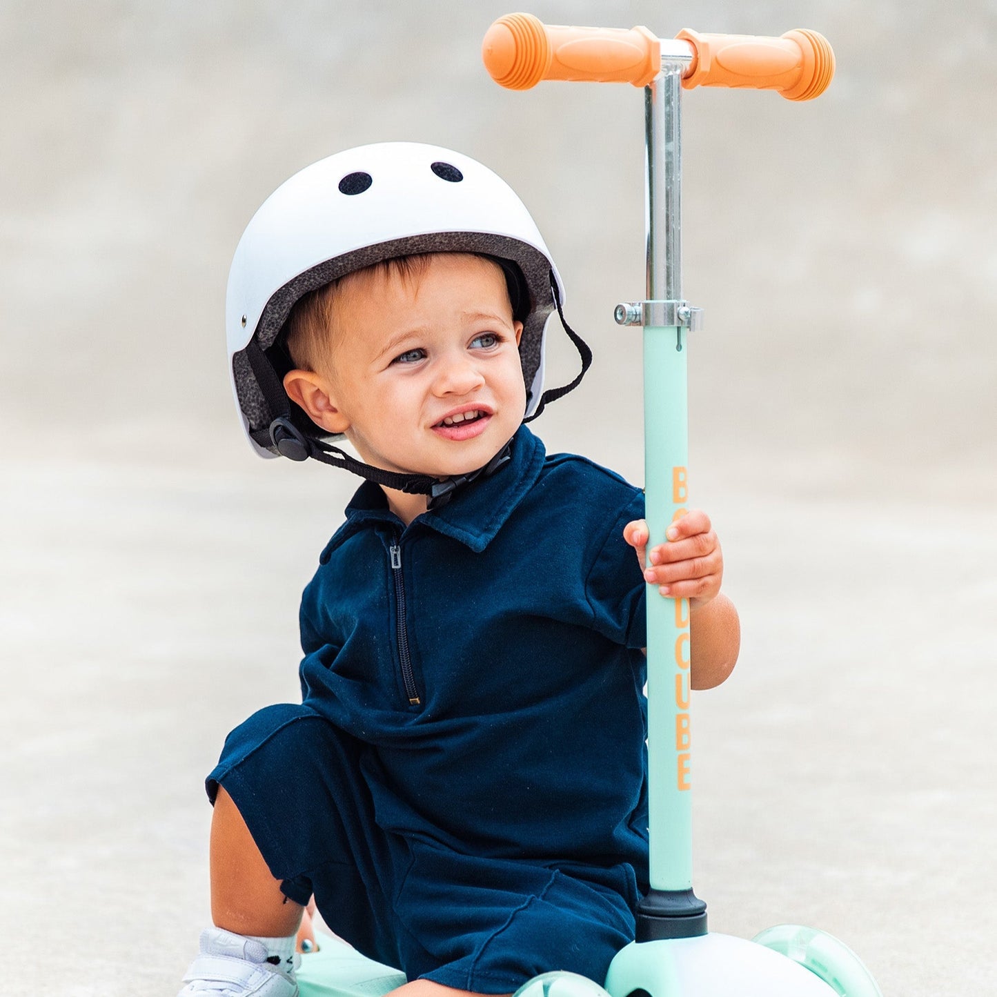 Child sitting on a scooter with a helmet, wearing a navy dress and white shoes.