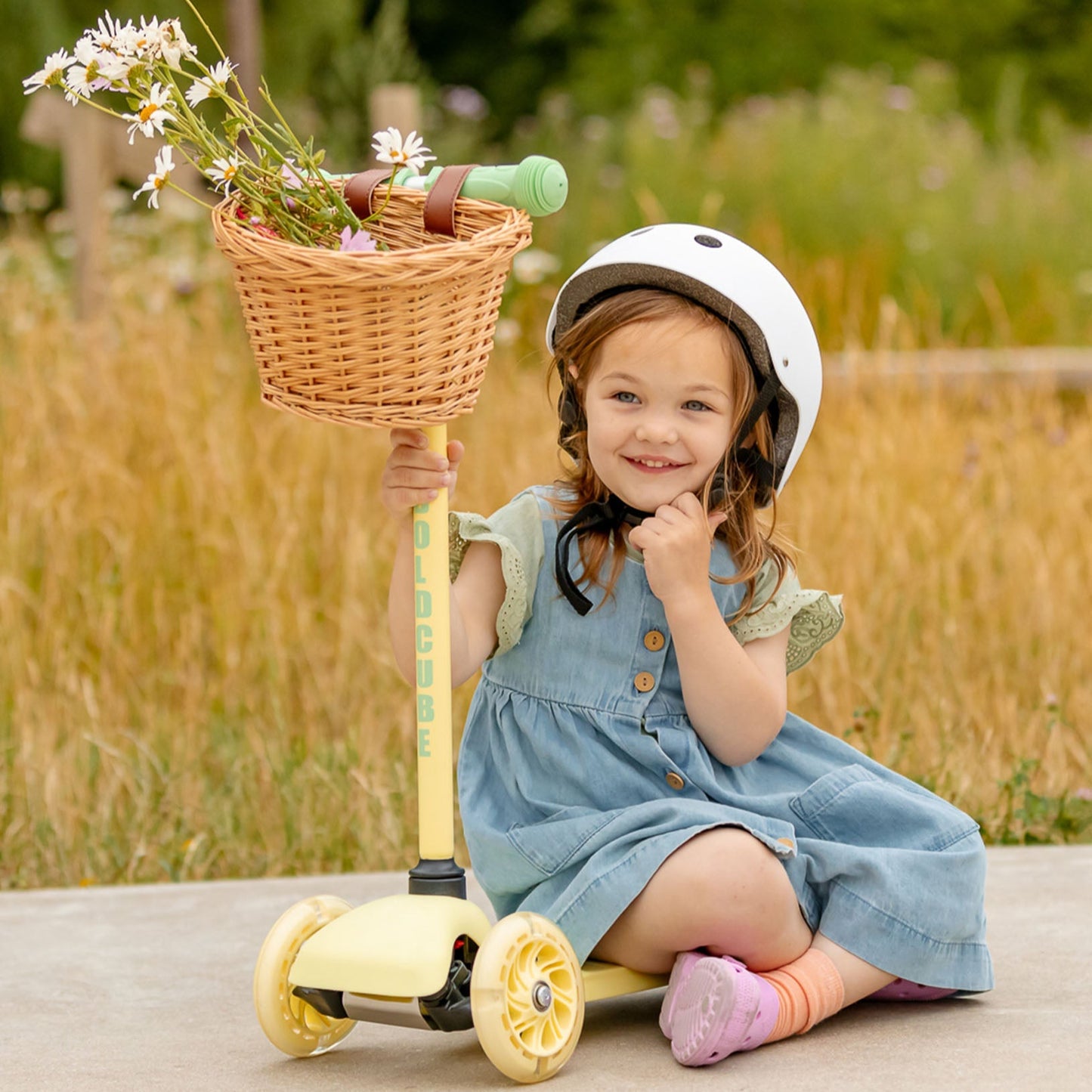 Child sitting on a scooter with a basket full of flowers, wearing a helmet in an outdoor setting.