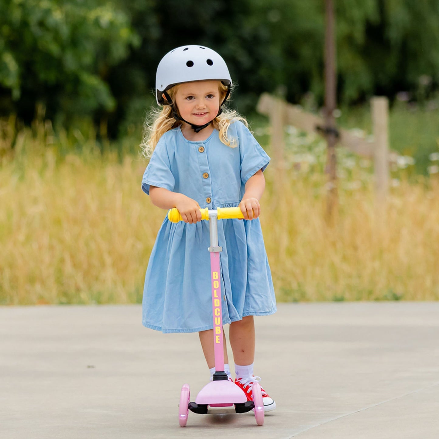 Child in a blue dress riding a pink scooter with a helmet on a path surrounded by greenery.
