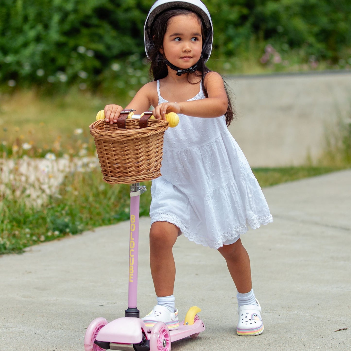 Child in a white dress standing on a scooter with a basket outdoors.