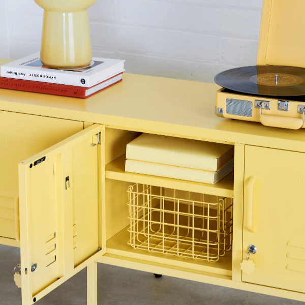 Yellow cabinet with open door displaying books and a wire basket, against a light gray wall.