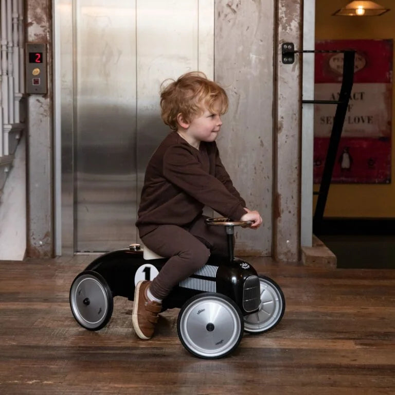 Child riding a toy car in an indoor setting with a metallic elevator in the background.