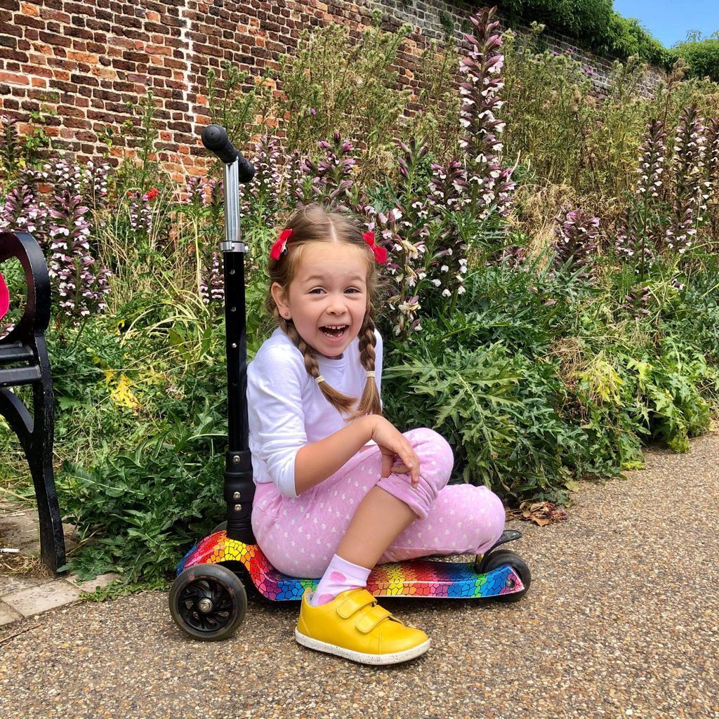 Young girl sitting on a scooter in a garden with flowers and a brick wall in the background