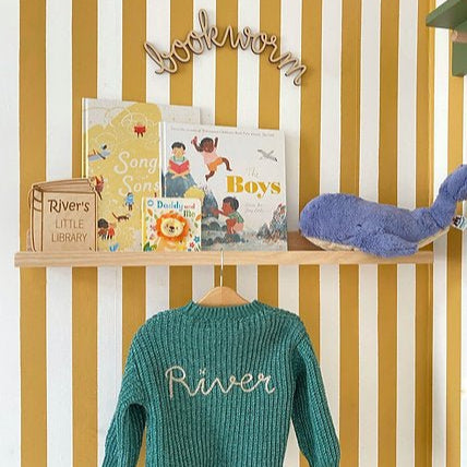 Children's sweater with 'River' embroidery, books, and a toy on a shelf against a striped wall.