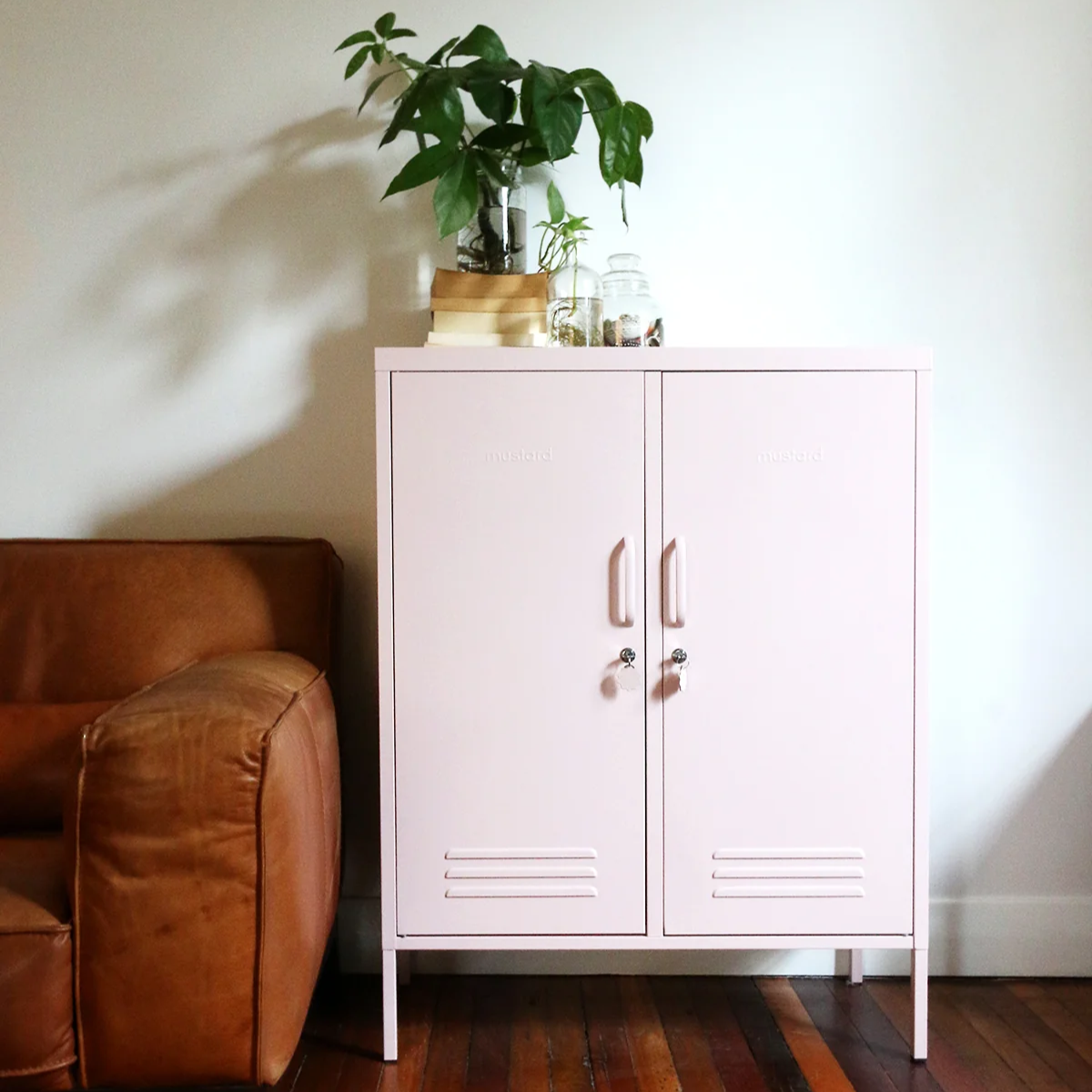 Pink cabinet with plants against a white wall, next to a brown leather chair on a wooden floor.
