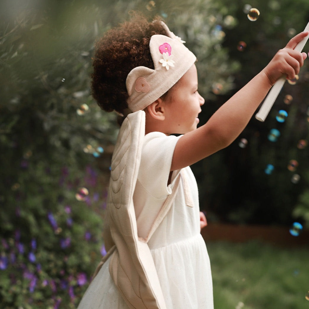 Child in a white dress with a headband, blowing bubbles outdoors.