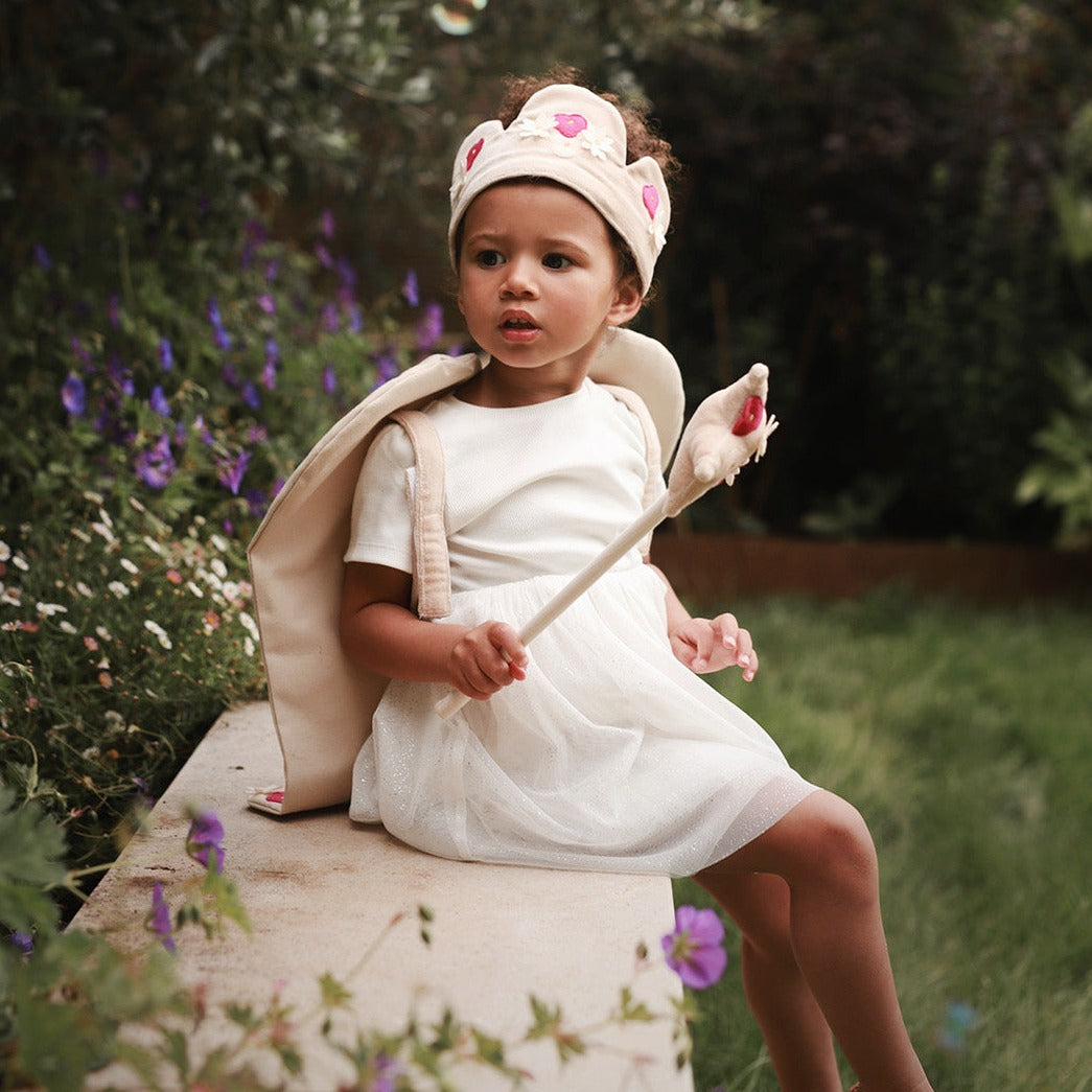 Child in a white dress with bunny ears and a wand, sitting outdoors with flowers in the background