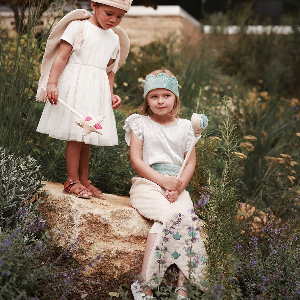 Two children in a garden setting, one standing and one sitting on a rock, holding toy swords.