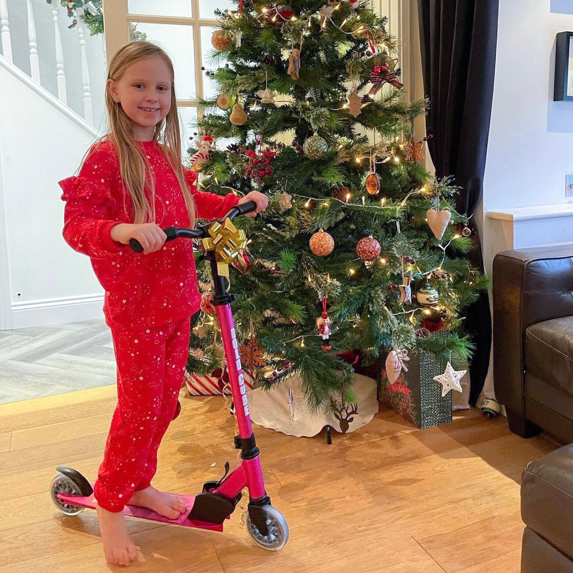 Child in red pajamas with a pink scooter in front of a decorated Christmas tree.
