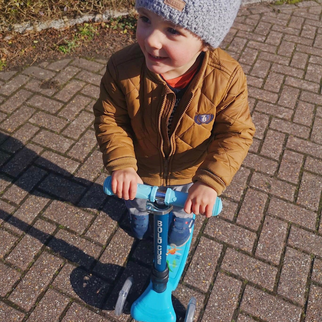Child on a blue scooter wearing a brown jacket and gray beanie on a paved surface.