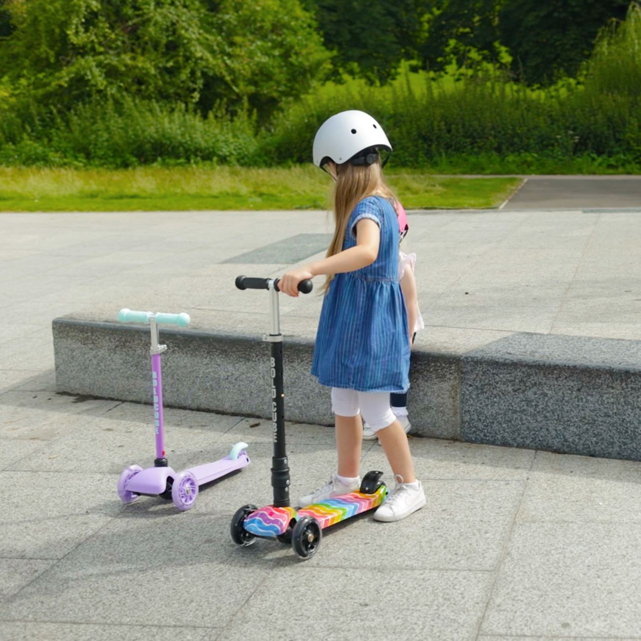 Child with a helmet standing next to a scooter on a paved area with greenery in the background