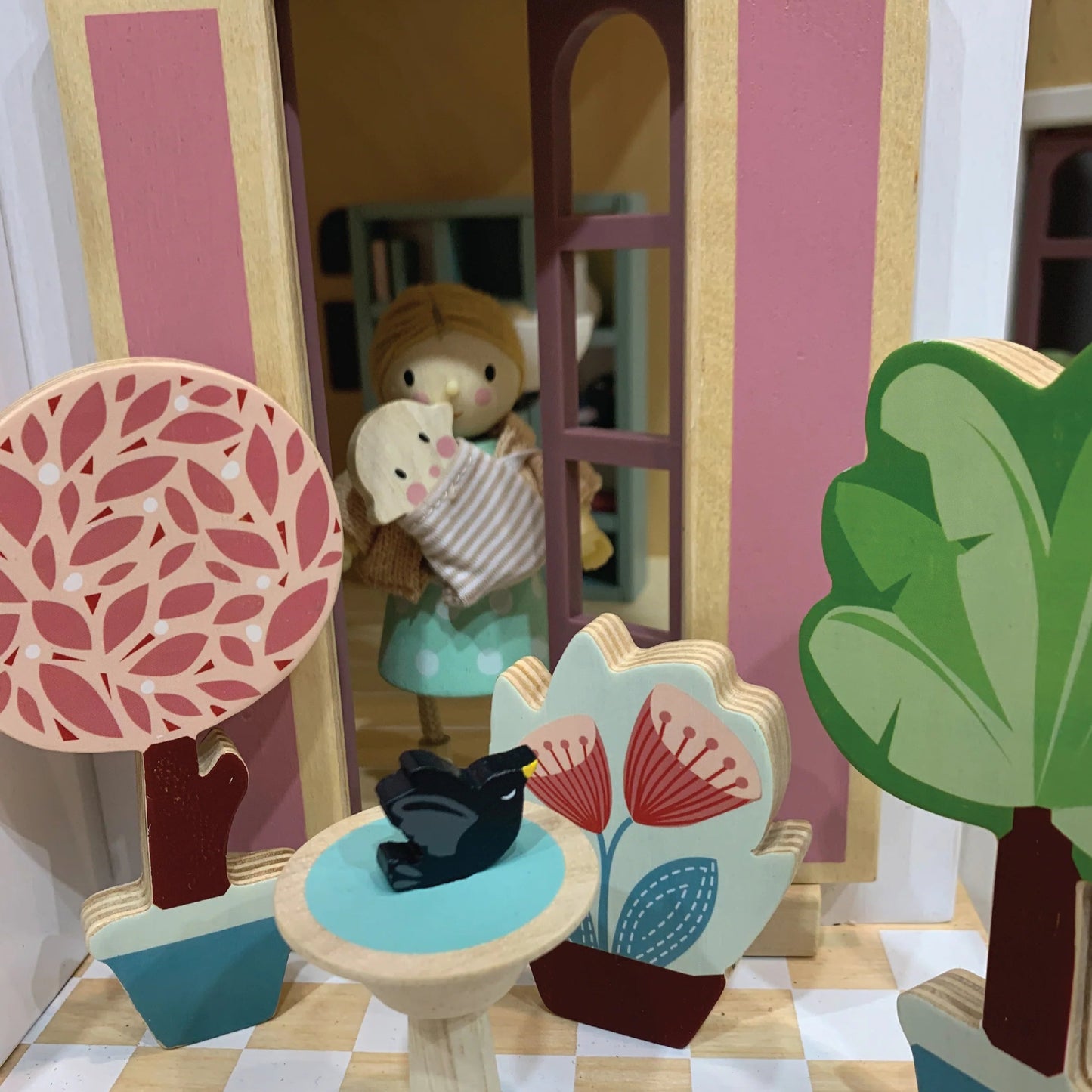 Toy house with a child figure holding a baby, surrounded by decorative trees and plants on a checkered floor.