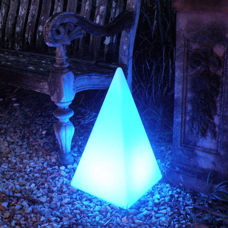 Pyramid-shaped LED lamp emitting a blue glow on a pebbled surface near a wooden bench.