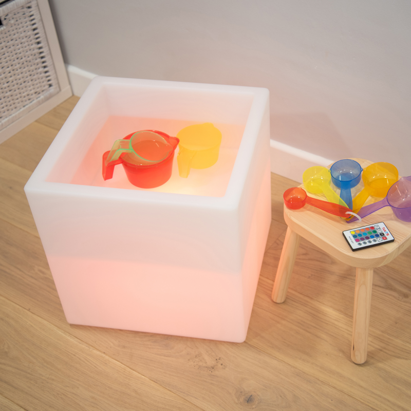 Colorful toys inside a glowing cube on a wooden floor with a small table and remote control.