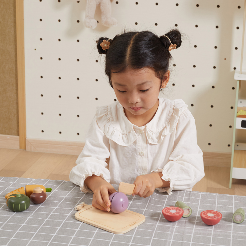 Young girl playing with wooden food toys on a checkered tablecloth.
