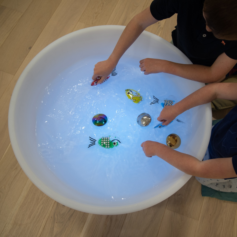 Children playing with colorful toys in a large water table filled with water on a wooden floor.