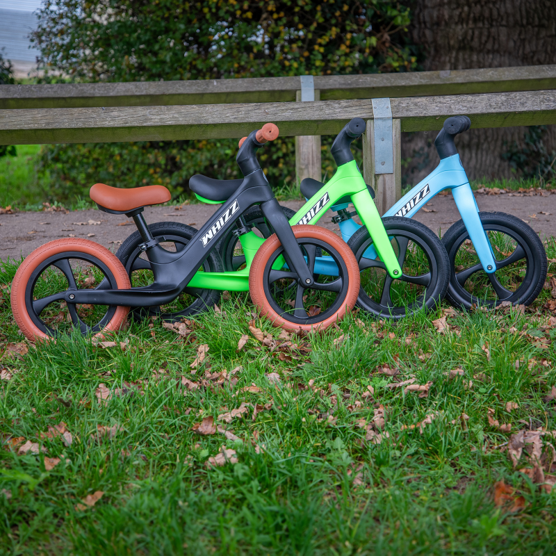 Three balance bikes in black, green, and blue on grass with a wooden fence in the background.