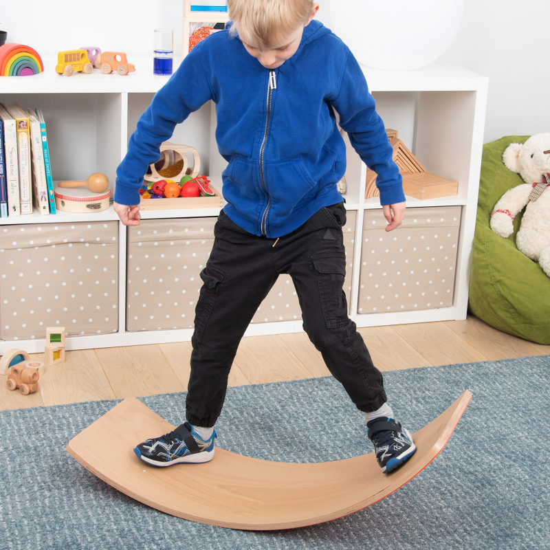 Child balancing on a wooden balance board in a room with toys and books.