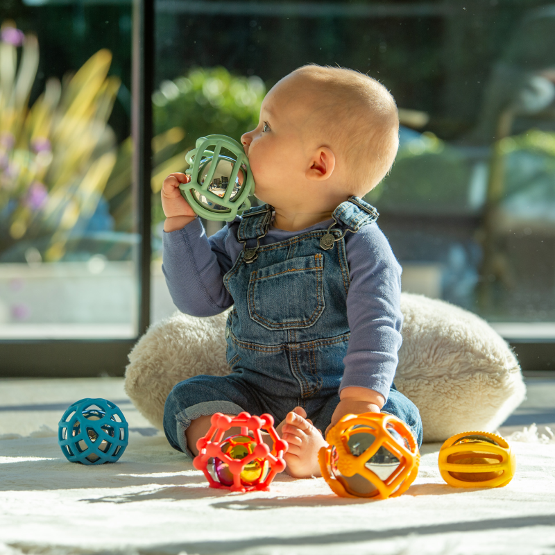 Baby playing with colourful silicone toys on a light surface, with a blurred outdoor background.