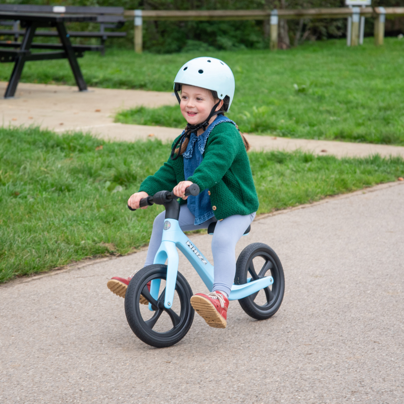 Child riding a blue balance bike on a sidewalk with grass and picnic tables in the background
