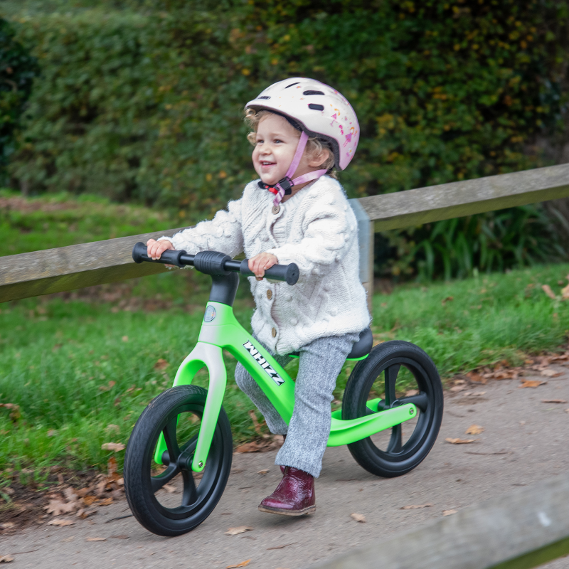 Child riding a green balance bike outdoors on a path with grass and trees in the background.