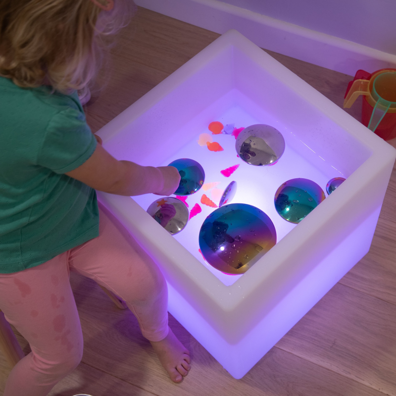 Child playing with colorful objects in a illuminated cube box on a wooden floor