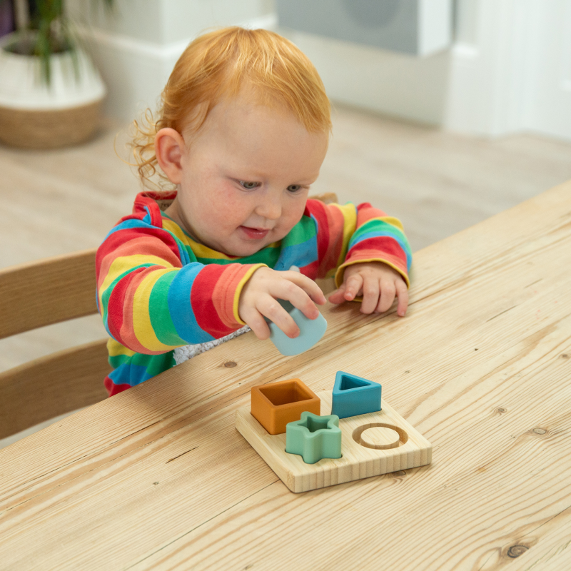 Child playing with a wooden and silicone shape sorting toy on a wooden table.