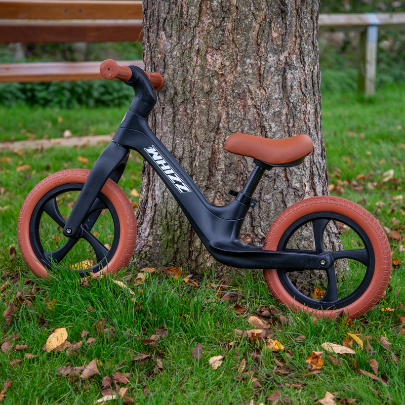 Black balance bike with brown wheels leaning against a tree in a park setting