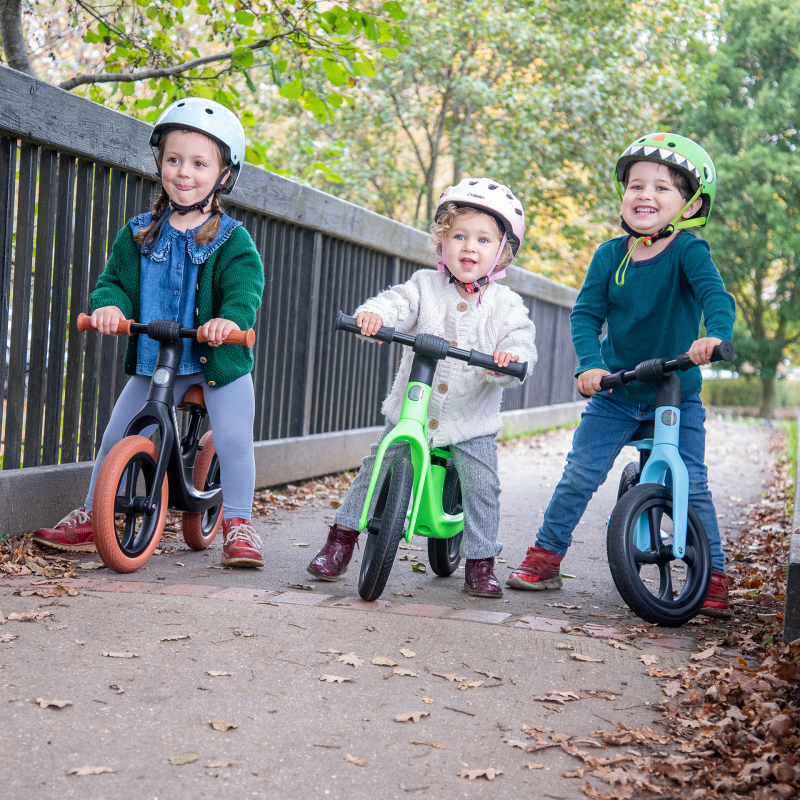 Three children riding balance bikes on a path with trees in the background