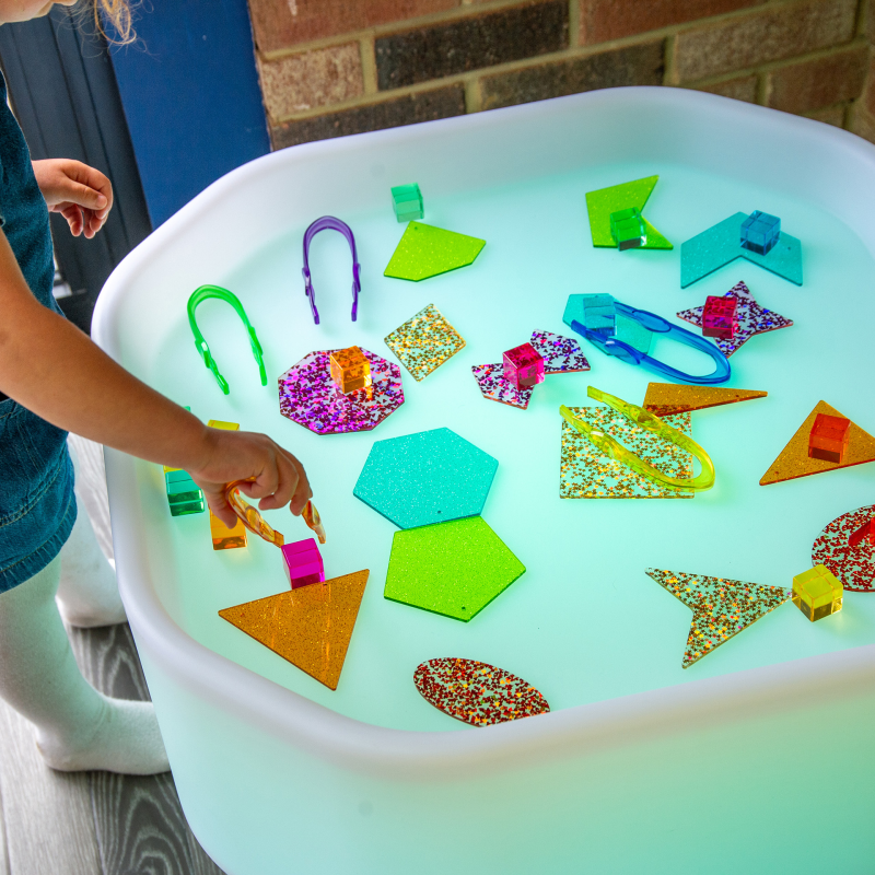 Children's play table with colorful geometric shapes and a child's hand reaching in.