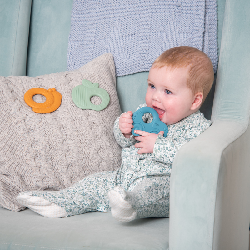 Baby sitting on a couch holding a blue teething ring with two additional teething rings on a pillow.