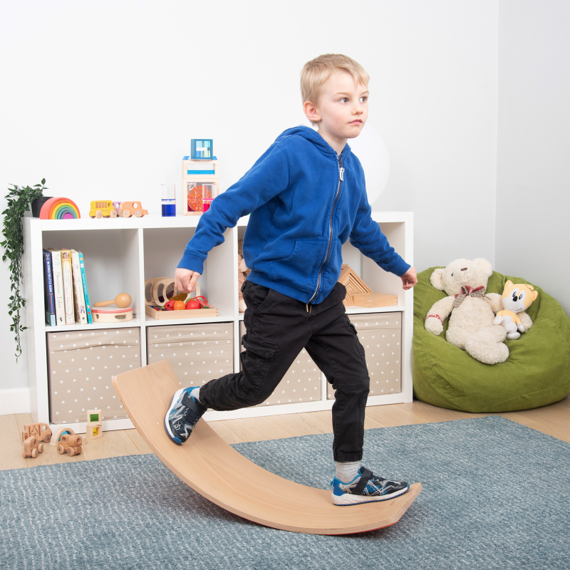 Child using a wooden balance board in a room with toys and books.