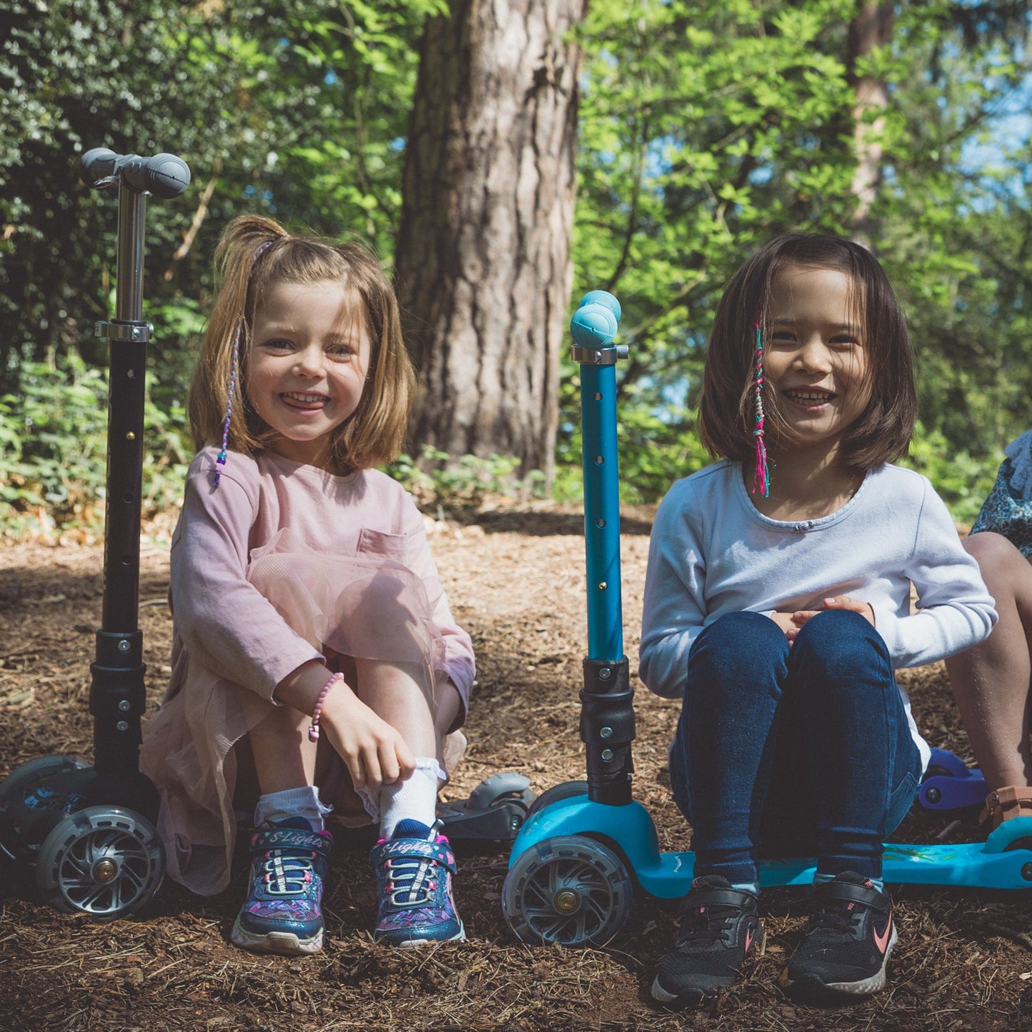 Two children sitting on scooters in a park with trees in the background