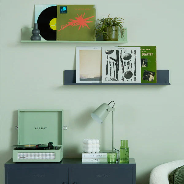 Vinyl record player on a cabinet with shelves displaying records and a lamp against a light green wall.