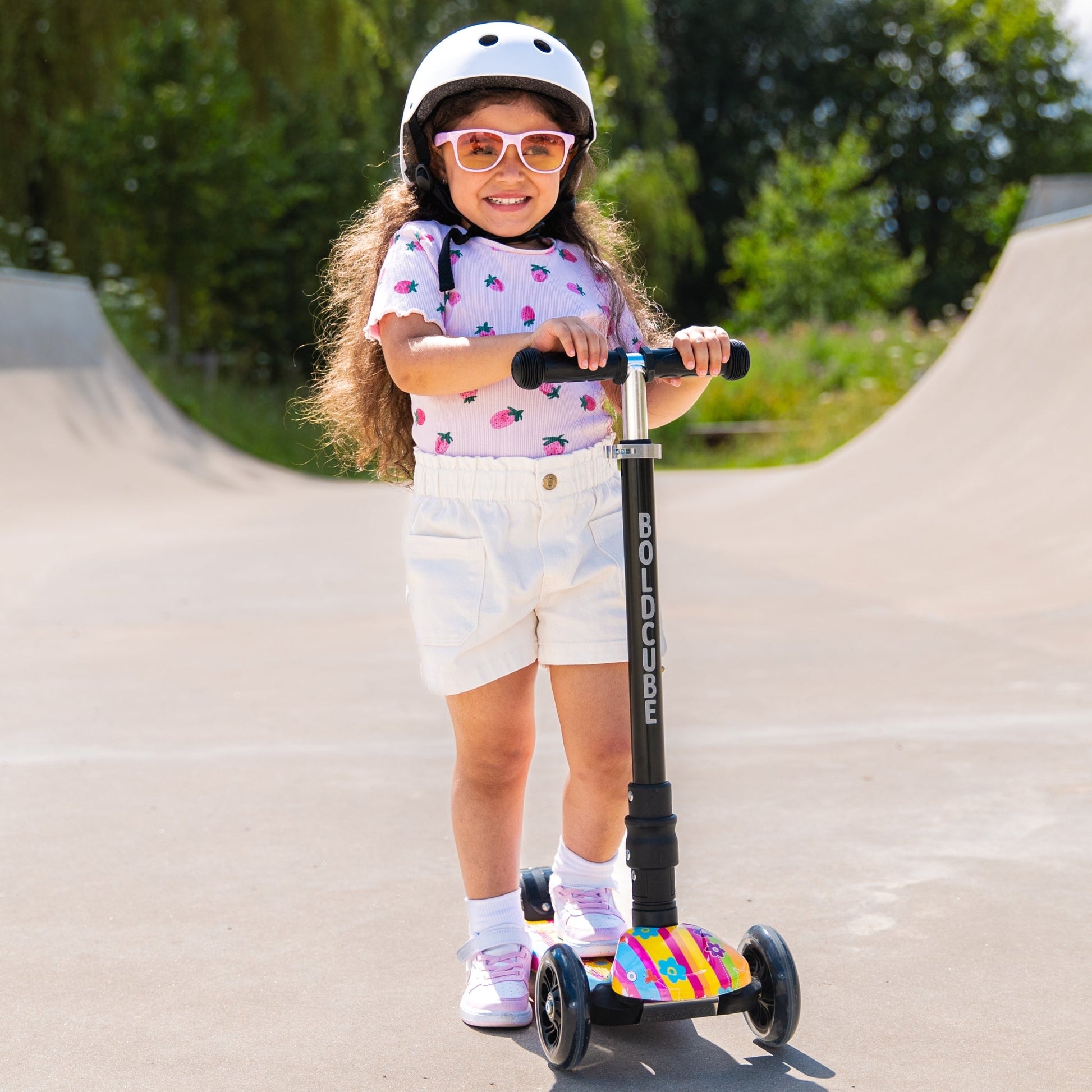 Child riding a scooter with a colorful deck on a paved path with greenery in the background