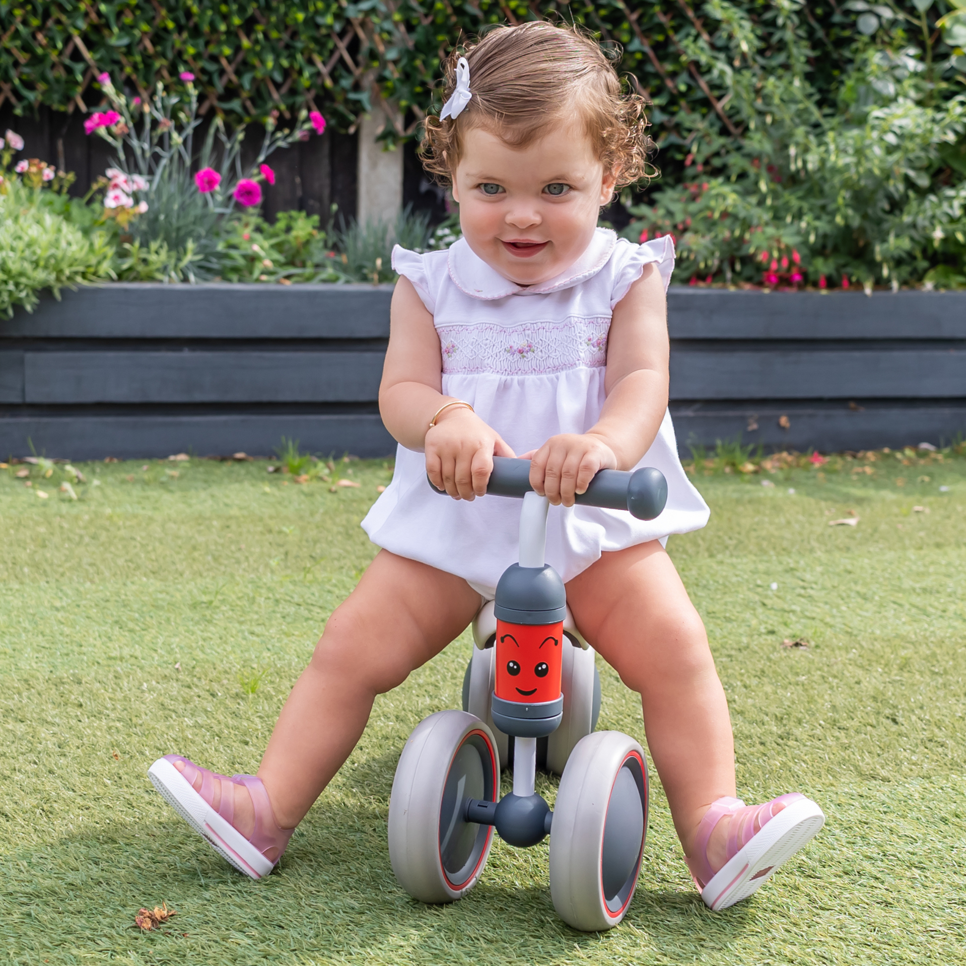 Child riding a toy bike outdoors on grass with flowers and plants in the background