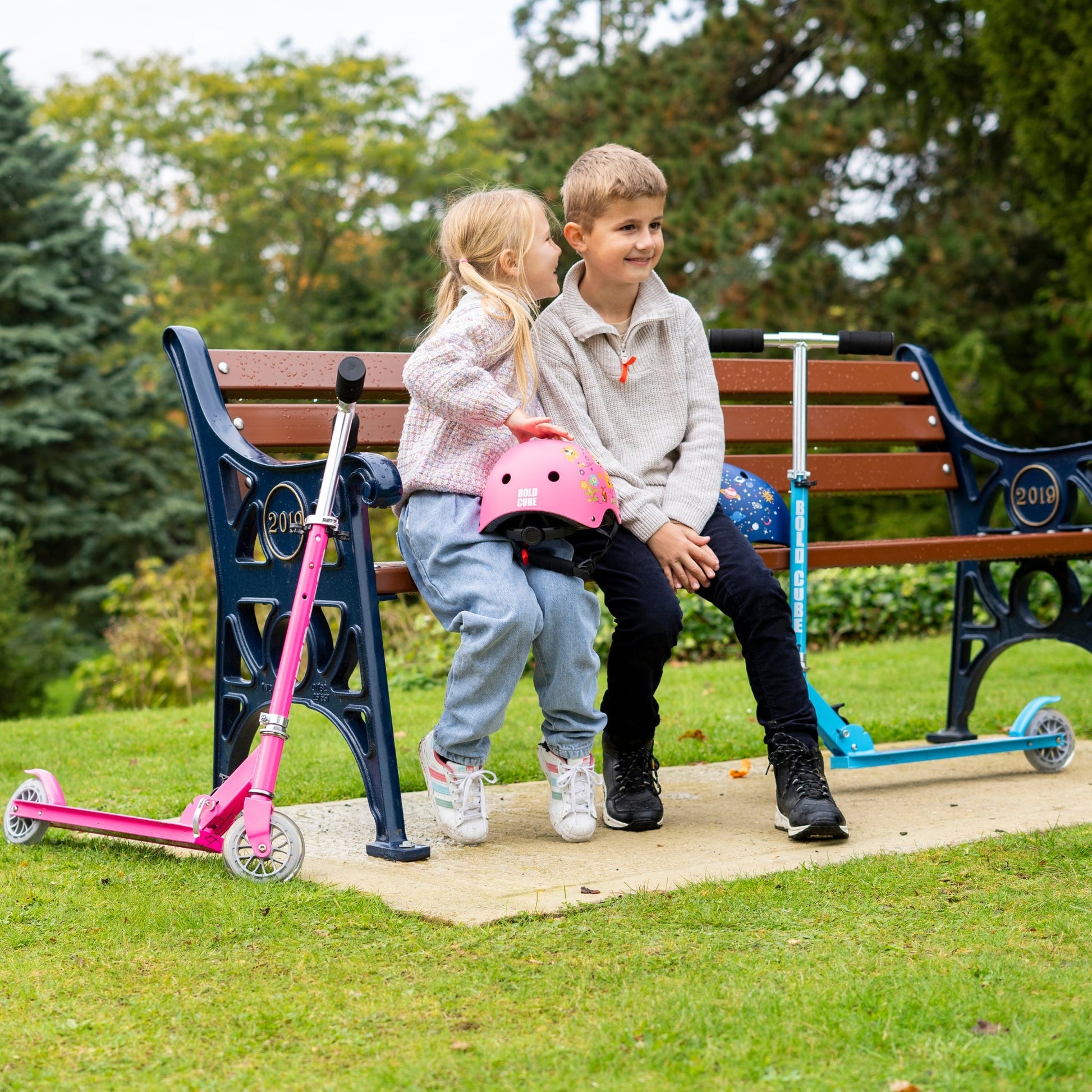 Two children with scooters sitting on a park bench