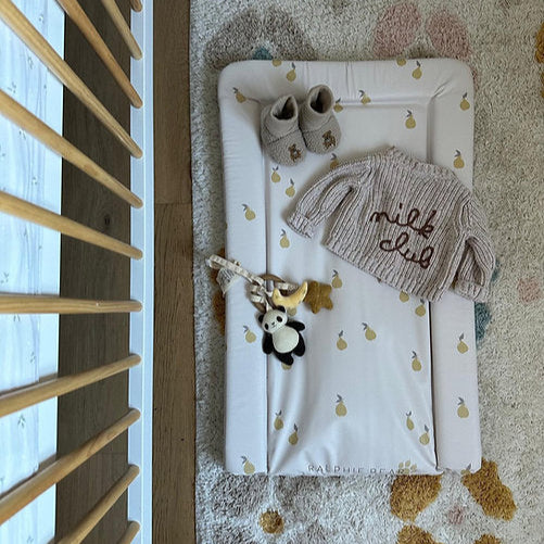 Baby hat and shoes on a changing table with toys, next to a wooden crib.