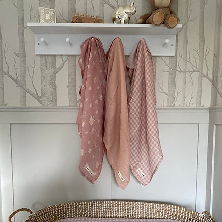 Pink baby blankets hanging on a shelf with a woven basket below against a tree-patterned wall.