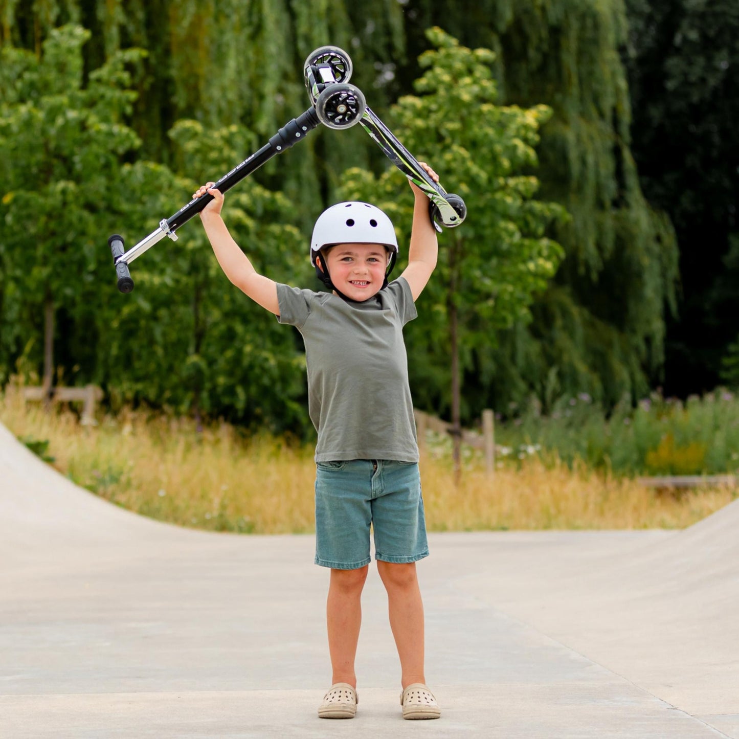 Child holding a scooter above their head with a helmet on, standing on a paved path with greenery in the background.
