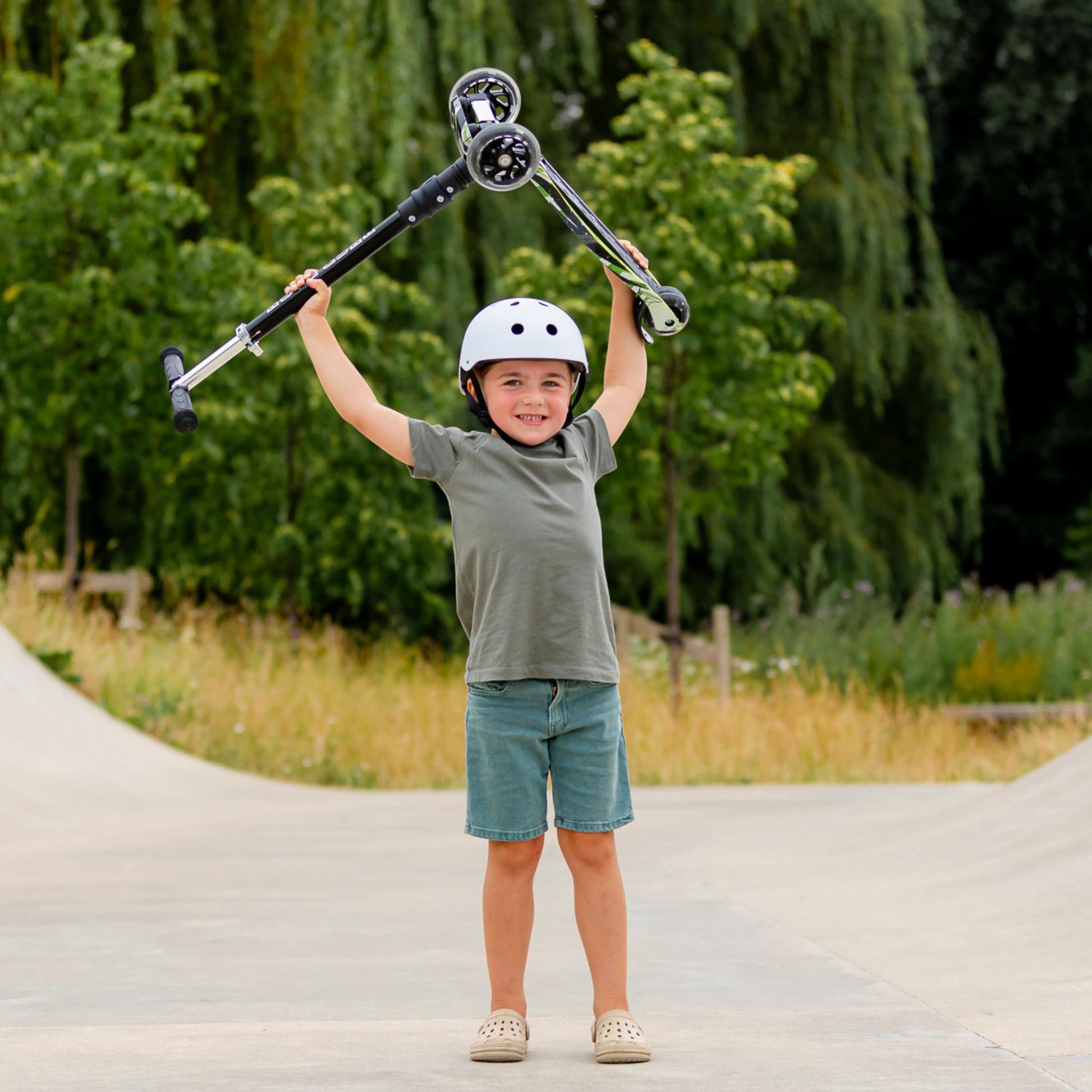 Child holding a scooter above their head with a helmet on, standing on a paved path with greenery in the background.