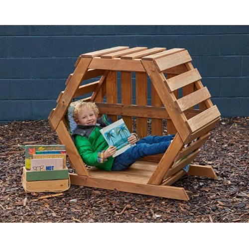 Child sitting in a wooden playhouse reading a book with books nearby on a mulched ground.