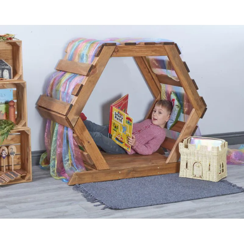 Child reading a book inside a wooden playhouse with colorful blanket and toys around.