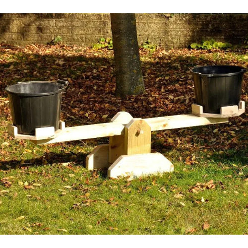 Wooden seesaw with two black buckets on a grassy area with leaves and a tree.