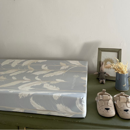 Baby crib mattress with feather pattern on a green surface next to baby shoes and a picture frame.