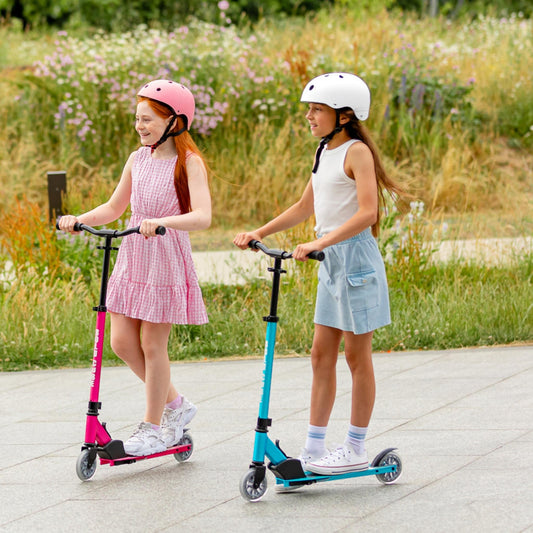 Two young girls riding scooters on a paved path with a grassy area and flowers in the background.